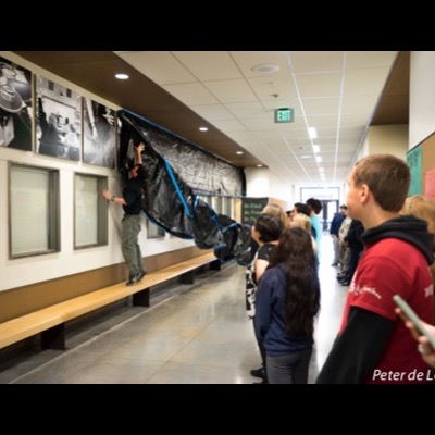 Photograph of the "A Pathway" hallway installation being hung at Hayes Freedom High School, a worker on a bench taping black protective plastic over the mounted photographs as students look on.