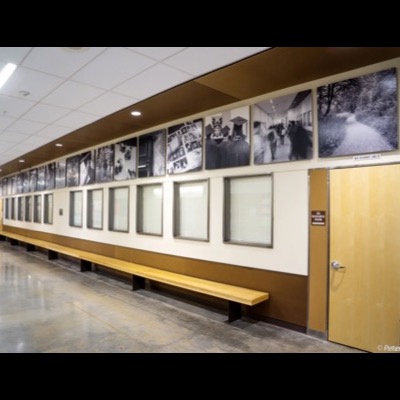 Photograph looking down the same school hallway at the installed black-and-white photograph series mounted above windows, a wooden bench below and a "132 Resource Room" door at the end.