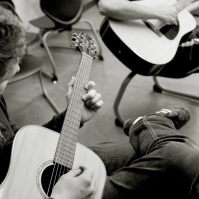Black-and-white photograph, from directly above, of a student’s hands fretting an acoustic guitar neck while seated in a classroom circle, another guitar-playing student visible behind.