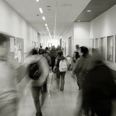 Black-and-white photograph looking down a busy school hallway, students in motion-blurred silhouette walking past a bulletin board and rows of windows.