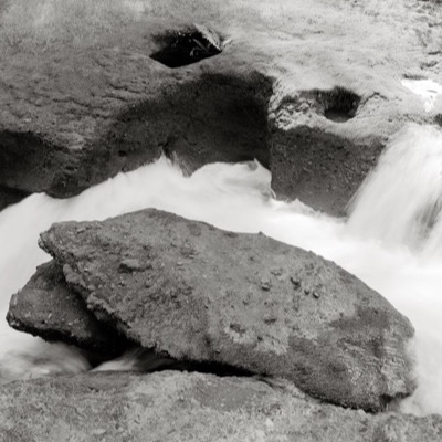 Black-and-white photograph of a large rock split by rushing whitewater, framed close against smaller boulders in the current.