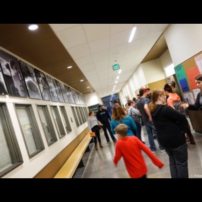 Photograph of visitors filling a school hallway beneath the installed "A Pathway" photographs, a woman in an orange jacket and others seated and standing near a "Be Kind" sign.