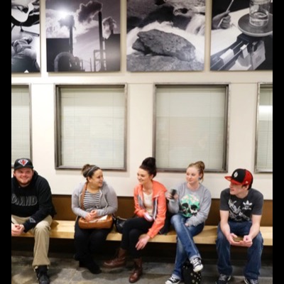 Photograph of five students sitting on a bench beneath four mounted black-and-white photographs — industrial smokestacks, a river boulder, and a potter’s hands at a wheel — talking and smiling together.
