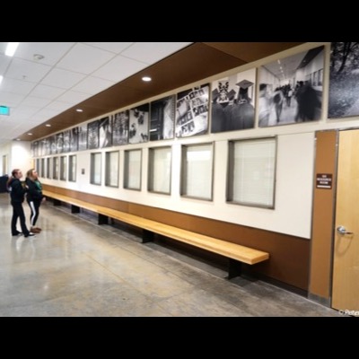 Photograph of two students looking up at the installed hallway photographs, including images of graduating students in caps and gowns and a blurred hallway crowd, near a "132 Resource Room" door.