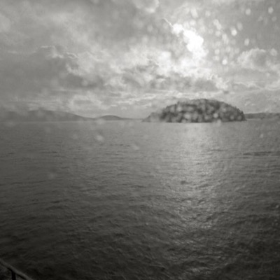 Black-and-white photograph, its surface flecked with water spots, of a forested island silhouette across the Rosario Strait, sunlight breaking through heavy clouds above the water and a ferry rail visible at the bottom edge.