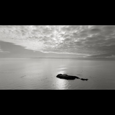 Black-and-white photograph of a calm sea beneath a heavily clouded sky, the sun’s glow diffused along a distant shoreline of low hills.