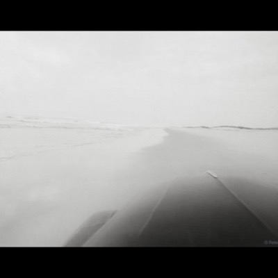 Black-and-white photograph, taken from a moving vehicle, of a hazy shoreline curving into the distance with gentle surf breaking on the left.