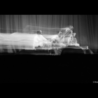 Black-and-white photograph, a long exposure, of a sitar player and two other musicians performing on stage, their forms streaked and blurred by motion against a heavy curtain backdrop.