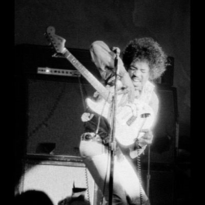 Black-and-white photograph of Jimi Hendrix performing live on stage, guitar raised and crossed over the microphone stand, caught mid-motion against a wall of Marshall amplifiers.