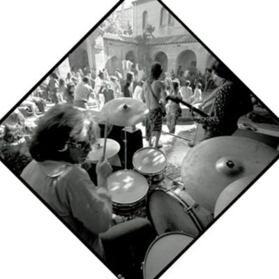 Black-and-white photograph, diamond-cropped, of a drummer performing at an outdoor courtyard concert, cymbals filling the foreground as a bassist and shirtless singer play to a crowd gathered under Spanish-style arches.