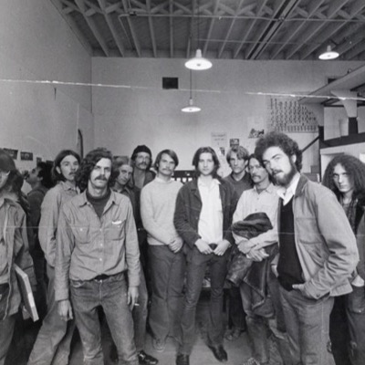 Black-and-white group portrait of about eleven young men standing together in a warehouse-like room with exposed rafters and hanging lamps, a mixing board and posters visible on the wall behind them.