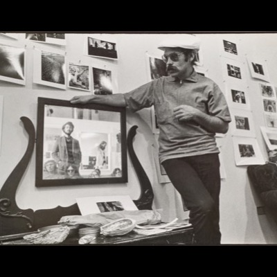 Black-and-white photograph of a young man in a newsboy cap and sunglasses standing beside a wall covered in tacked-up photographs, resting his hand on a large framed group portrait.