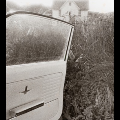Black-and-white photograph of an open car door beside a wire fence and overgrown grass, a farmhouse visible in the background across the field.