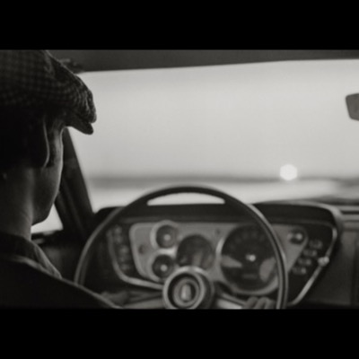 Black-and-white photograph, from the passenger seat, of a driver in a flat cap steering at dusk, dashboard gauges and the windshield glowing with fading light.