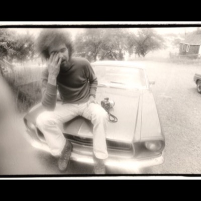 Black-and-white photograph of a bearded man with a large afro sitting on the hood of a vintage Ford Mustang, rubbing his eye, a twin-lens camera resting on the hood beside him.