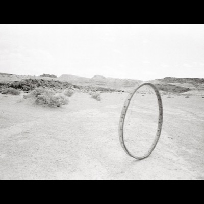 Black-and-white photograph of a bicycle wheel rim standing upright alone in an empty desert wash, low scrubby hills and sparse brush in the background.