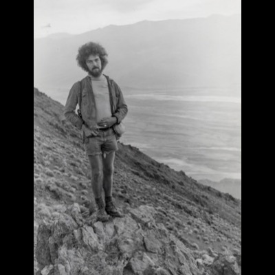 Black-and-white photograph of a bearded young man with a large afro in cut-off shorts standing on a rocky outcrop overlooking a vast desert valley and distant mountains, likely Death Valley.