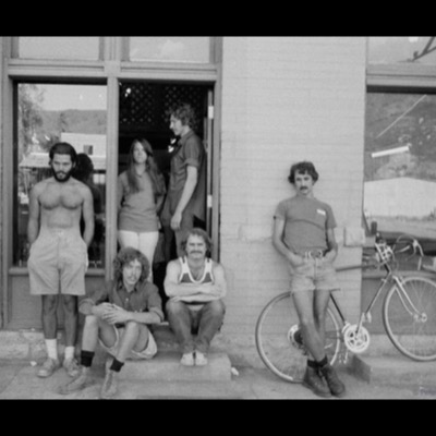Black-and-white photograph of six young men and one woman posed outside the Center of the Eye Gallery storefront, one shirtless, another leaning against a bicycle propped against the brick wall.