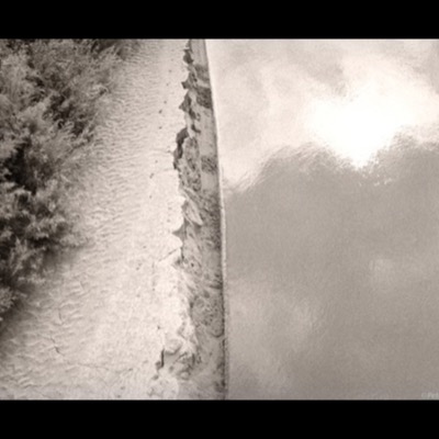 Black-and-white photograph of a rippled sandy riverbank cut by an eroded seam of dark earth, brush lining the bank on the left and glare-lit water filling the right half of the frame.