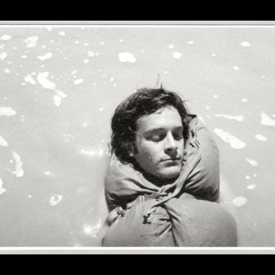 Black-and-white close-up portrait of a young man asleep with his head resting against a bundled sleeping bag, wet snow visible melting on the ground around him.