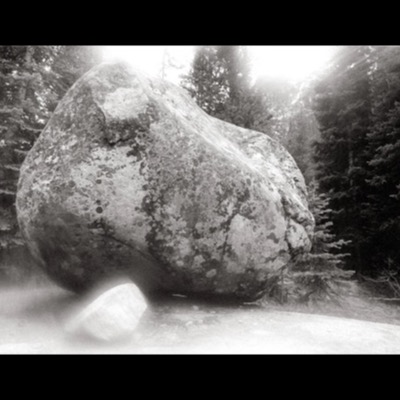 Black-and-white photograph of a large, lichen-covered glacial boulder balanced on a smaller rock in a conifer forest clearing, backlit by bright sunlight breaking through the trees.