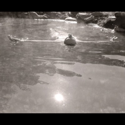 Black-and-white photograph of a nude figure floating on their back in a rock-lined hot spring, arms outstretched, sunlight reflecting brightly off the water.