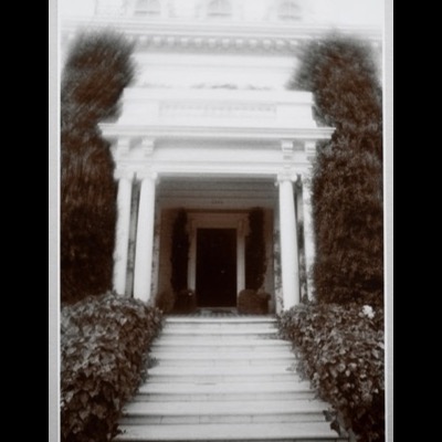 Black-and-white photograph of a grand columned entrance portico with a wide staircase flanked by ivy hedges, the numbers "2590" visible above the dark doorway.