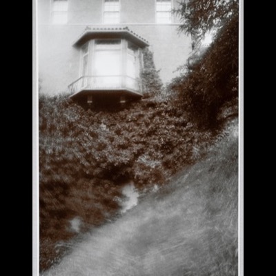 Black-and-white photograph of an octagonal bay window and balcony on the mansion’s upper floor, framed by dense ivy climbing the wall below.