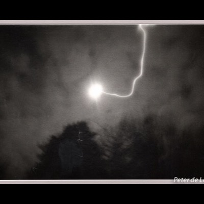 Black-and-white photograph of a bright light source in a night sky with a glowing, branching streak arcing across dark storm clouds above silhouetted treetops.