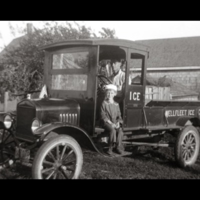Color archival photograph of a young boy in a sailor cap sitting on the running board of a vintage Ford ice-delivery truck marked "ICE" and "Wellfleet Ice Co.," a driver at the wheel above him.