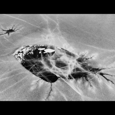 Black-and-white photograph, an abstract aerial view of cracked lake ice, a dark open patch of water breaking through a web of surface fractures.