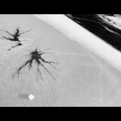 Black-and-white photograph, an abstract aerial view of ice, two dark branching cracks radiating across a granular frozen surface beside a smooth dark band.