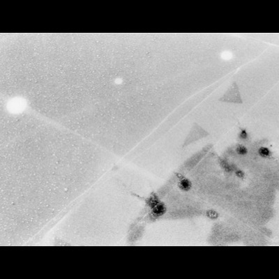 Black-and-white photograph, grainy and high-contrast, of light reflections and dark spots scattered across a snow-dusted icy surface, ridged snowdrifts crossing the lower right.