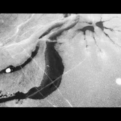 Black-and-white photograph, an abstract aerial view of dark open water curving between granular ice banks, with fine radiating cracks branching across the upper right.