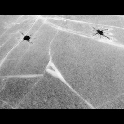Black-and-white photograph, an abstract view of two dark, spider-like cracks radiating symmetrically across a frost-covered ice surface.