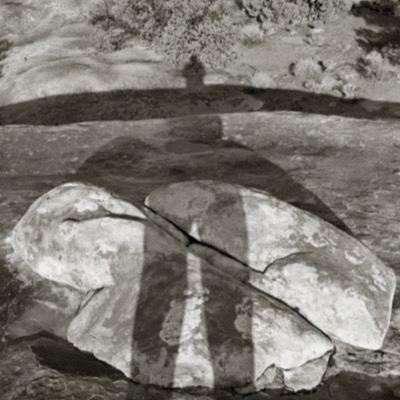 Black-and-white photograph looking down at two lichen-covered boulders split by a crack, a hiker’s shadow with a walking pole cast across them on a red-rock slickrock desert.
