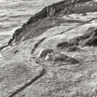 Black-and-white photograph looking down a grassy, wind-bent coastal bluff to a rocky point, ocean surf visible below and a worn footpath curving along the edge.