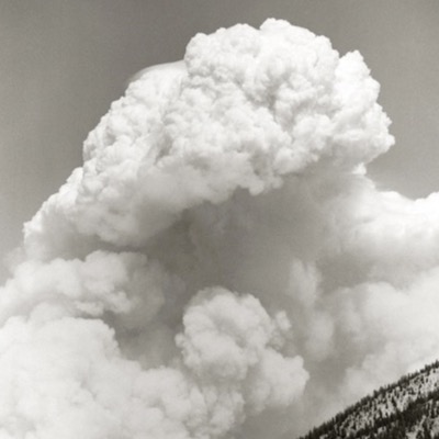 Black-and-white photograph, "Idaho Fire Cloud," of a massive cumulus tower billowing upward, dark storm base below and a snow-dusted forested ridge at the bottom edge.