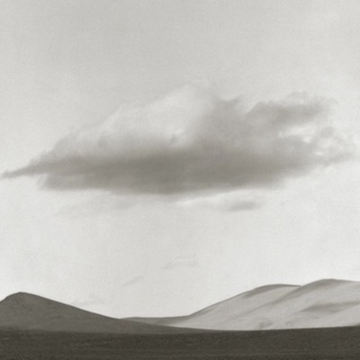 Black-and-white photograph of a solitary elongated cloud floating low over dark desert hills and dunes.