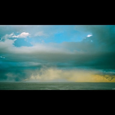 Color photograph, "Black Rock," of a hazy storm front moving over a flat desert playa, teal-tinted sky darkening from blue to near-black with sunlit mountains glowing beneath it.