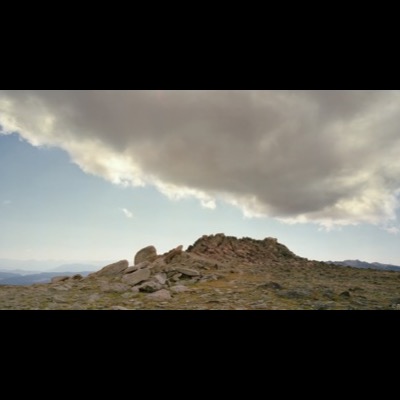 Color photograph of a rugged rock cairn on a mountain summit beneath a long, dark storm cloud stretching across a clear blue sky, distant peaks visible on the horizon.