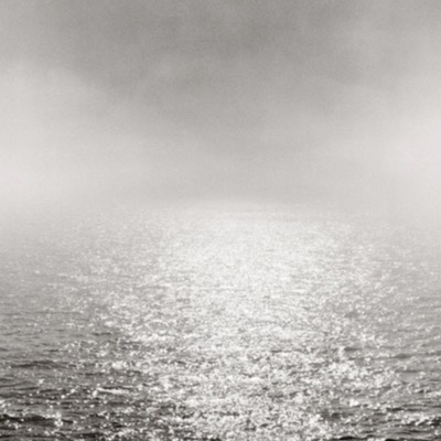 Black-and-white photograph of a bright, foggy sea horizon, the sun’s glare diffused into a soft glow over rippling water.