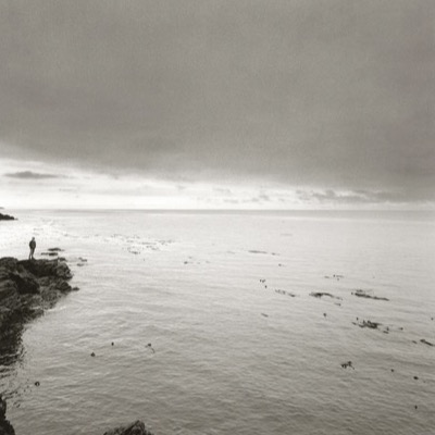 Black-and-white photograph of a lone figure standing on a rocky point looking out over a calm bay scattered with floating kelp, heavy clouds pressing low over the water.
