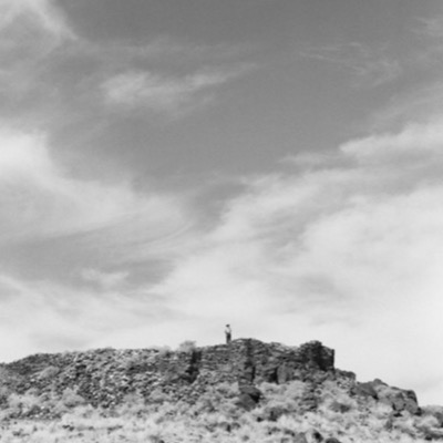 Black-and-white photograph of a lone figure in a hat standing atop a ruined stone wall on a hilltop, a wide cloud-streaked sky filling most of the frame above.