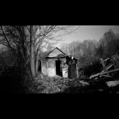 Black-and-white photograph of a collapsing wooden shed engulfed by bare trees at night, star trails streaking faintly across the sky and wreckage scattered beside it.