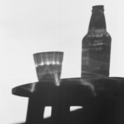 Black-and-white photograph, softly blurred, of a drinking glass and beer bottle balanced on the rounded back of a dark chair or table against a plain wall.