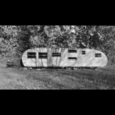 Black-and-white photograph of an abandoned, windowless vintage travel trailer with broken windows, parked in tall grass at a forest edge, roofing panels stacked beside it.