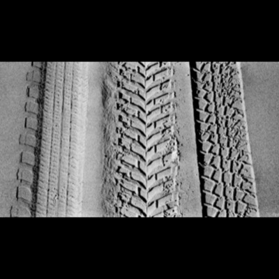 Black-and-white close-up photograph of three sets of tire tracks pressed into fine sand, their treads sharply defined by raking light.