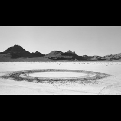 Black-and-white photograph of a large circular tire-track ring etched into a dry desert lakebed, jagged mountain peaks rising along the horizon behind it.
