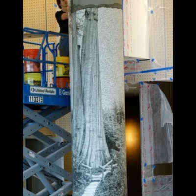 Photograph of the column mid-installation on a construction lift, the mosaic tree partly applied above bare cement and plastic sheeting protecting the surrounding wall.